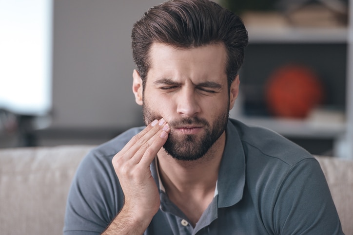 Frustrated young man touching his cheek and keeping eyes closed while sitting on the couch at home